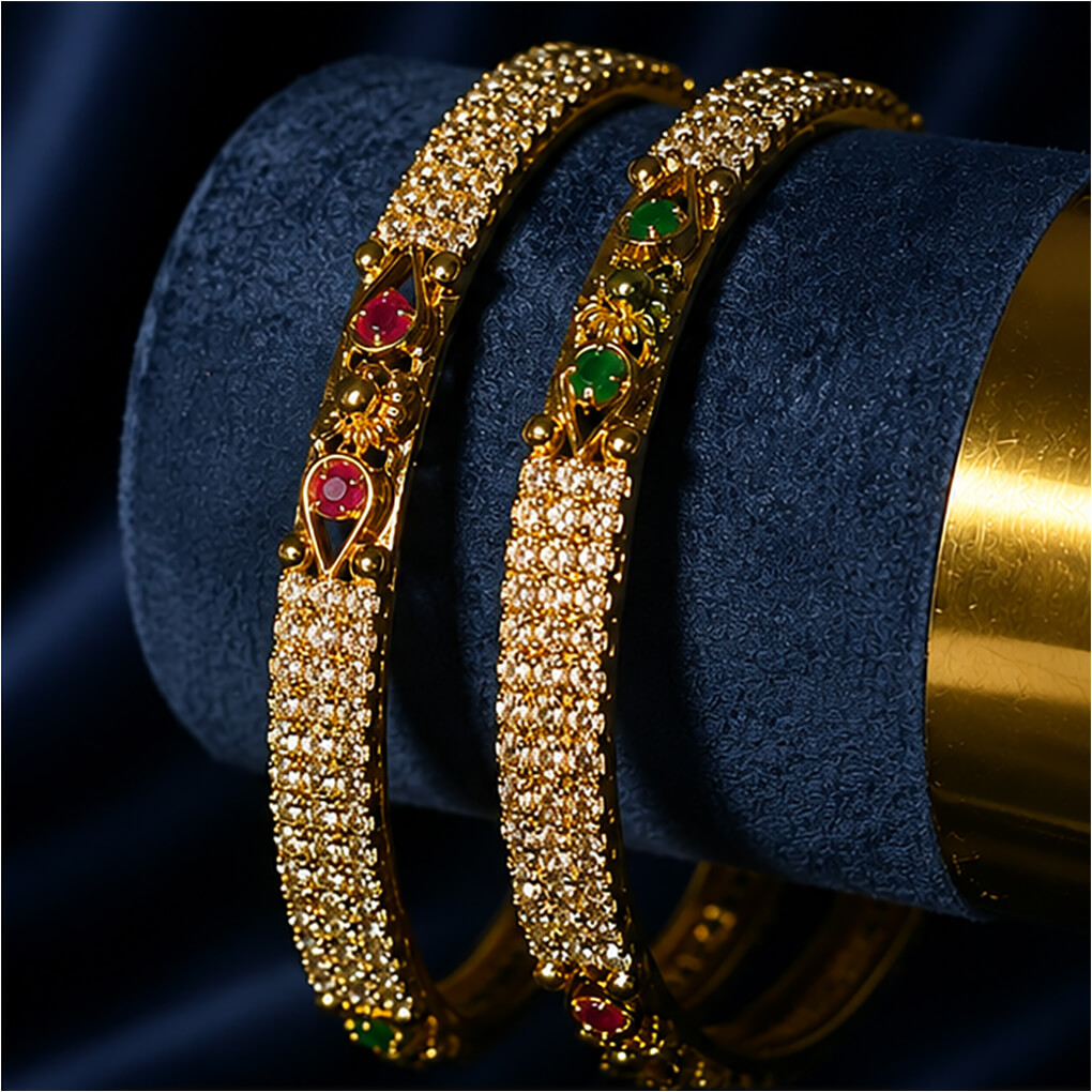 A pair of gold-tone crystal-studded bangles featuring emerald-green and ruby-red stone accents displayed on a luxurious navy blue background.