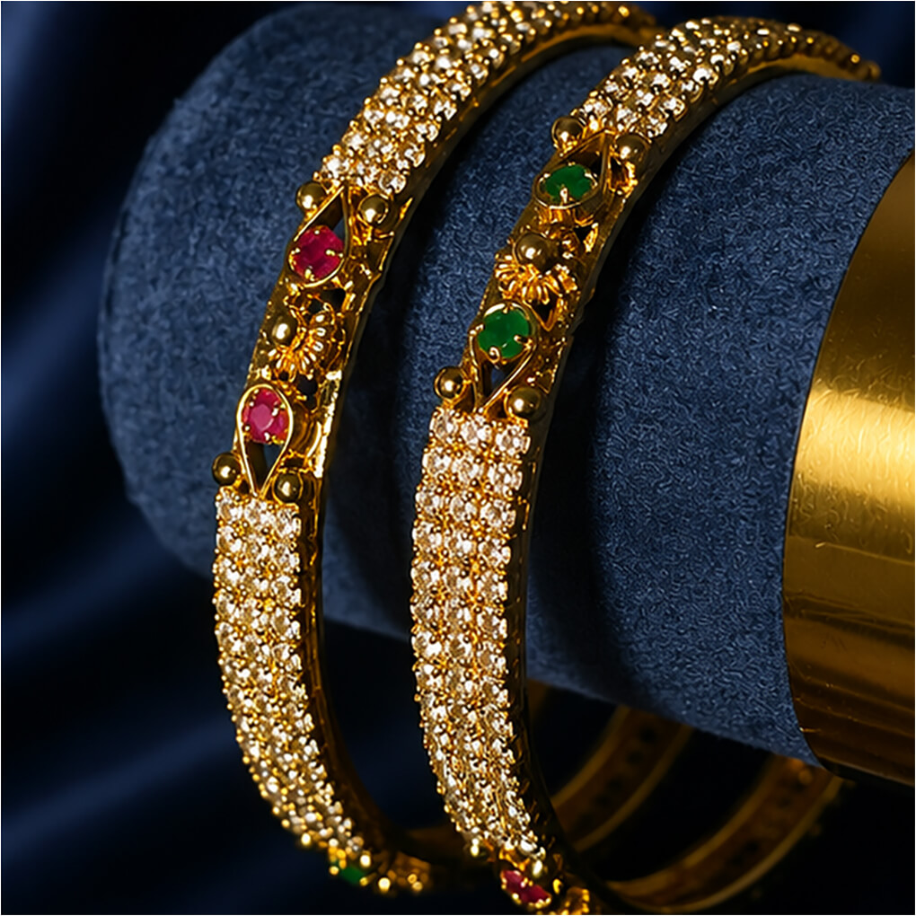 A pair of gold-tone crystal-studded bangles featuring emerald-green and ruby-red stone accents displayed on a luxurious navy blue background.