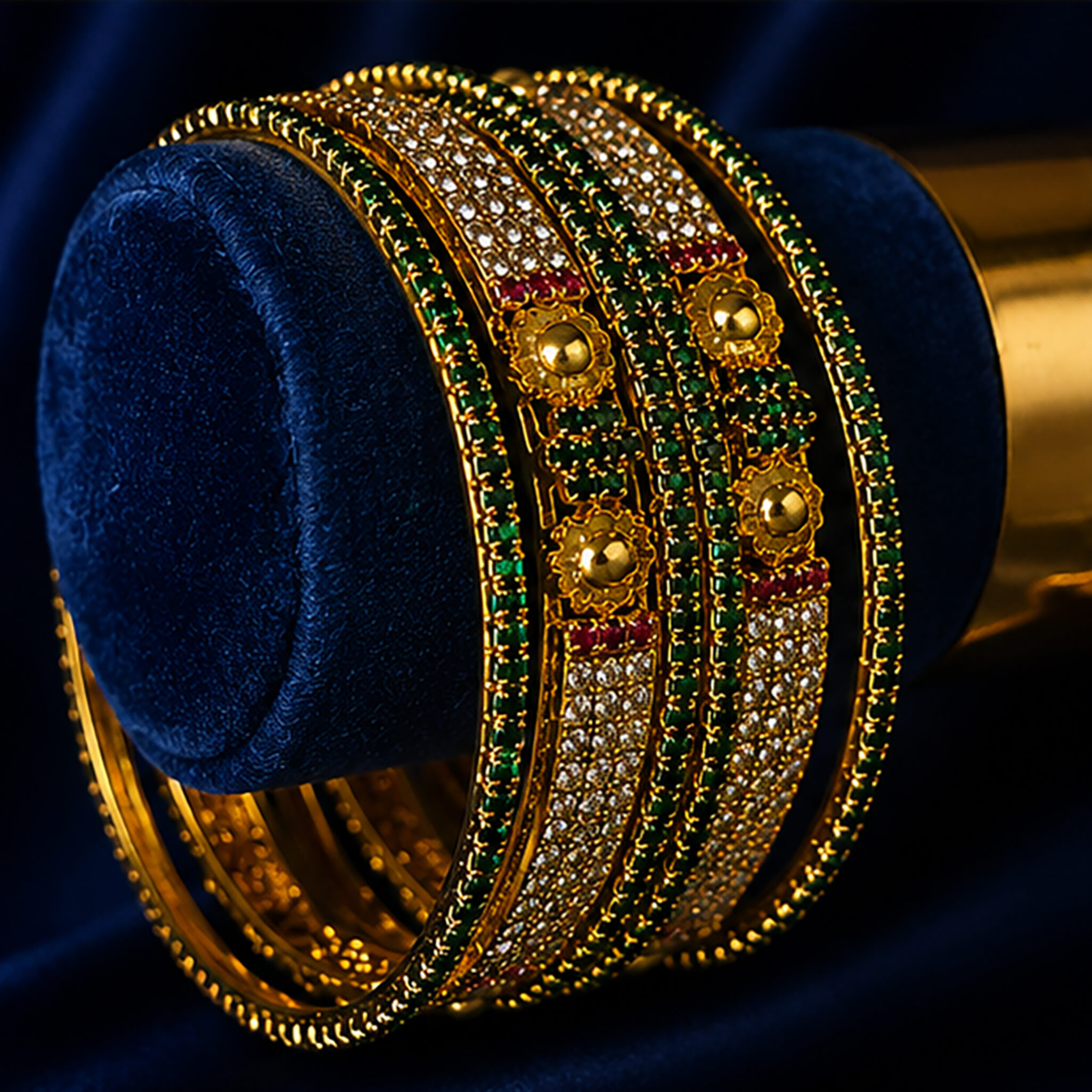 Gold-tone bridal bangles set featuring emerald-green stones, crystal embellishments, ruby accents, and floral motifs on a navy blue background.