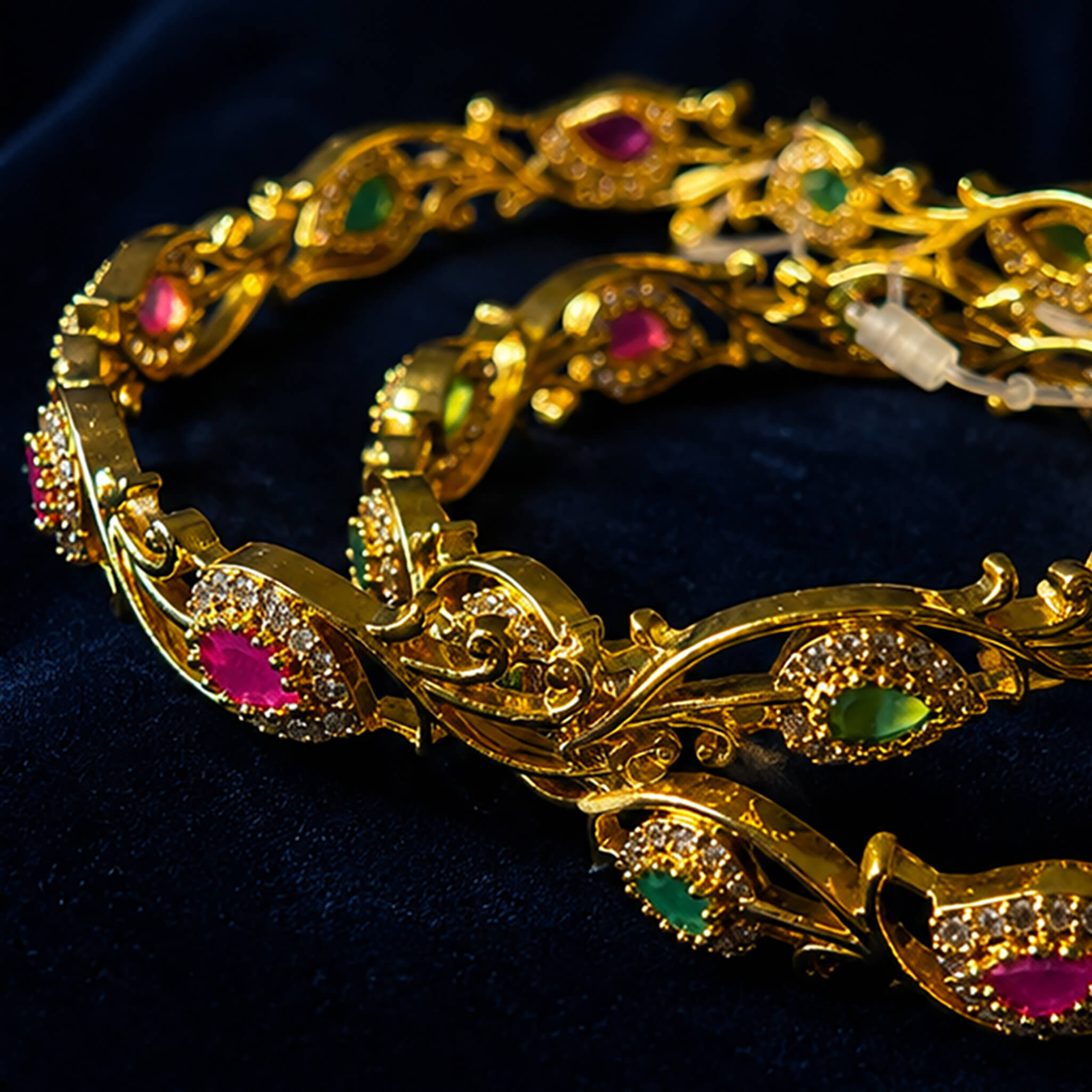 Gold-tone vine-pattern bangles featuring emerald-green and ruby-red stones with crystal embellishments on a navy blue background.