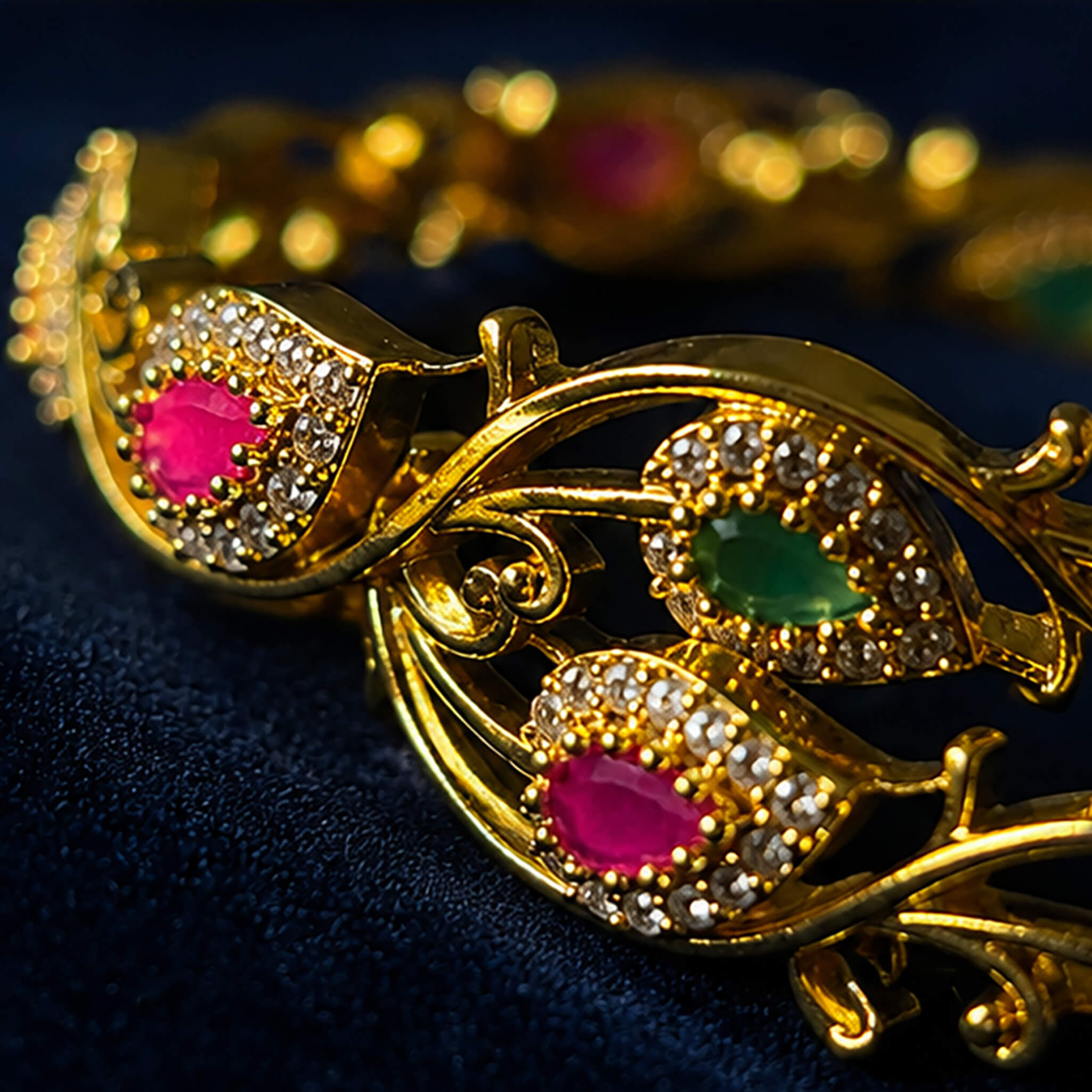 Gold-tone vine-pattern bangles featuring emerald-green and ruby-red stones with crystal embellishments on a navy blue background.