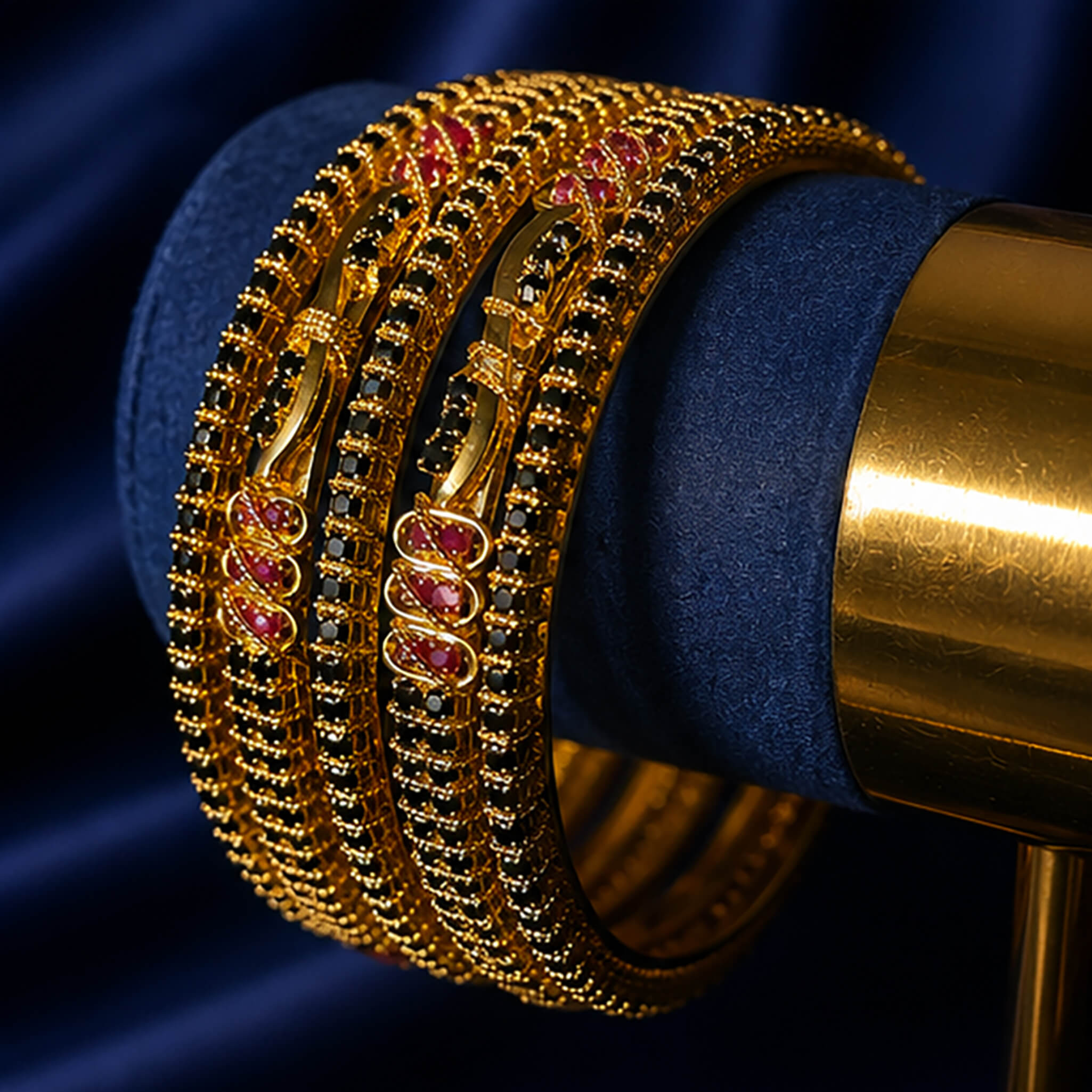 Gold-tone bangles featuring black stone detailing and ruby-red accents displayed on a luxurious navy blue background.