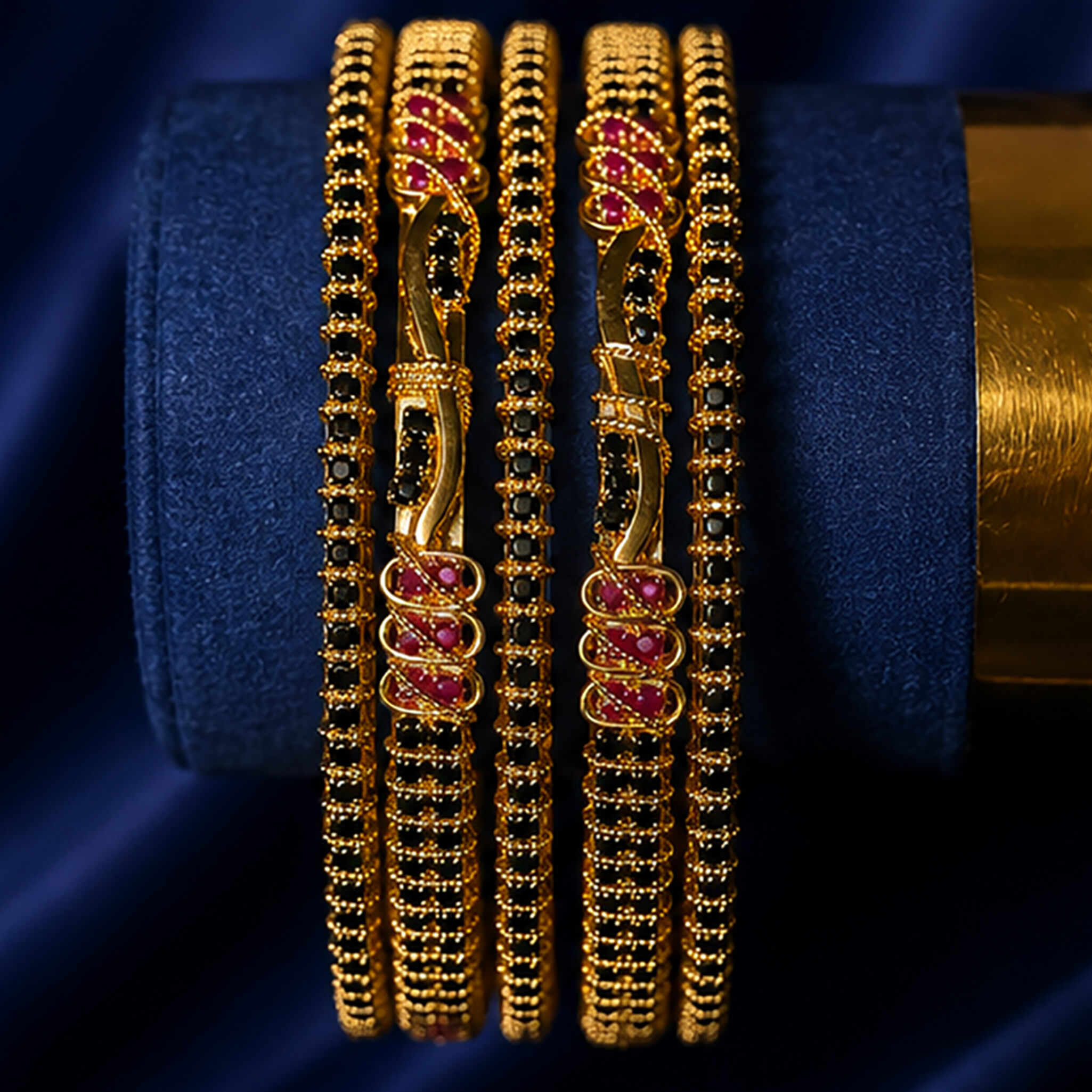 Gold-tone bangles featuring black stone detailing and ruby-red accents displayed on a luxurious navy blue background.