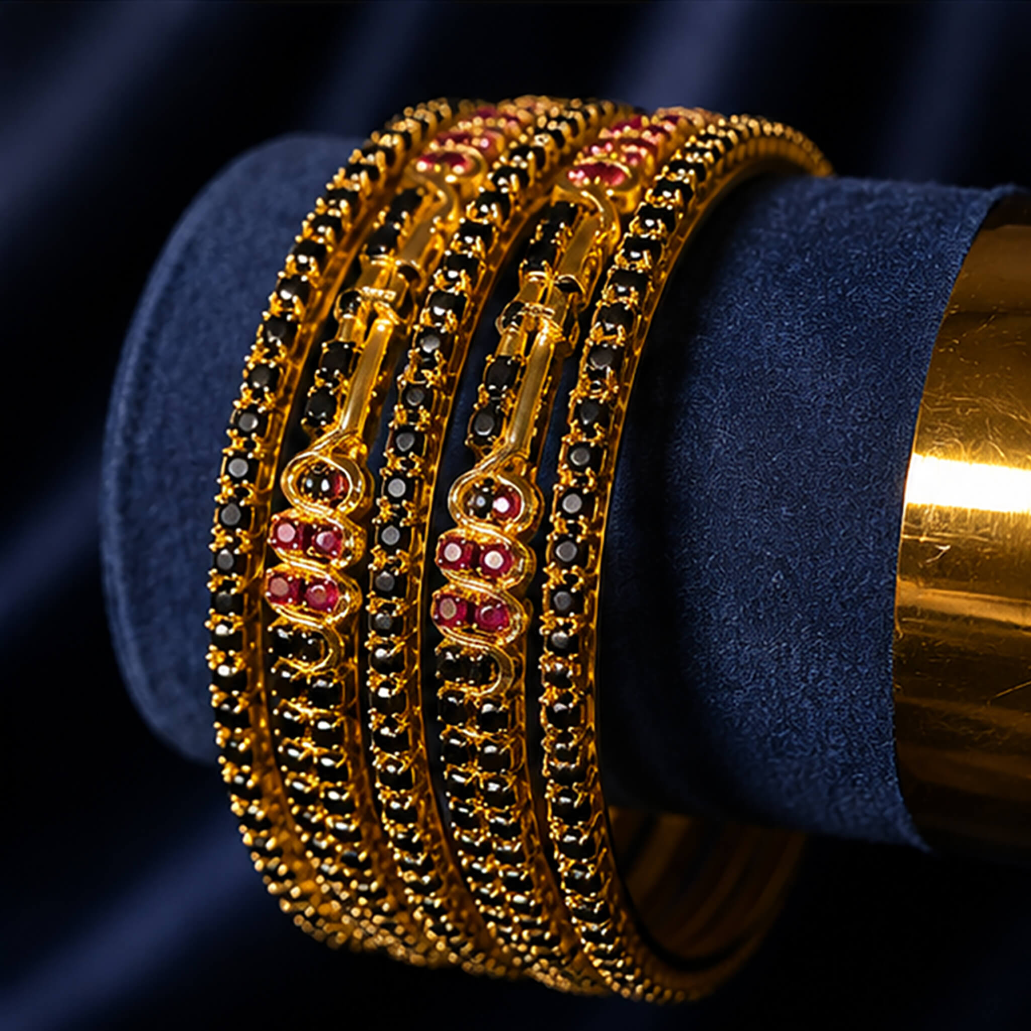 Gold-tone bangles with black stone embellishments and ruby-red accents displayed on a navy blue velvet background.