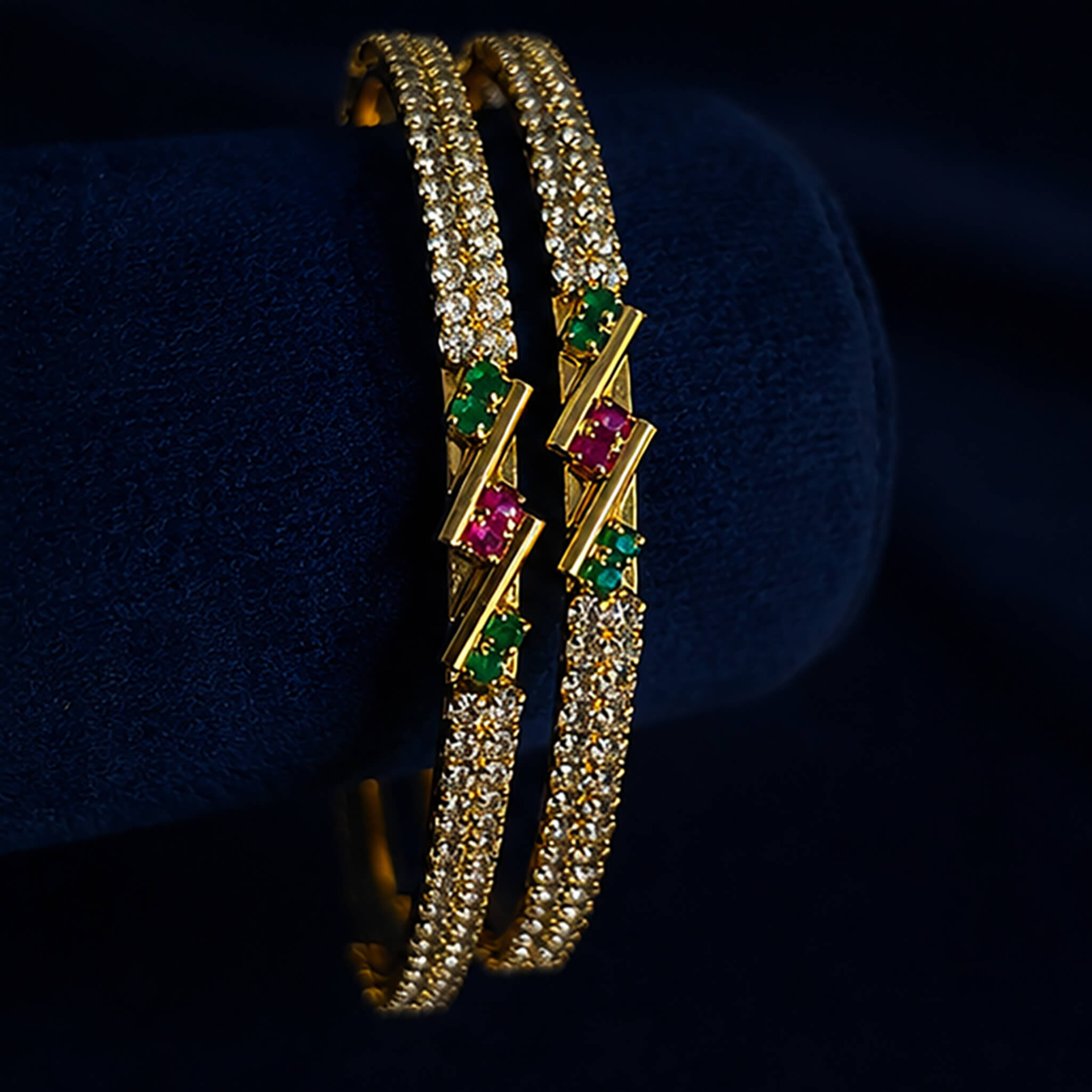 Gold-tone crystal-studded bangles with ruby-pink and emerald-green accents displayed on a blue velvet stand.