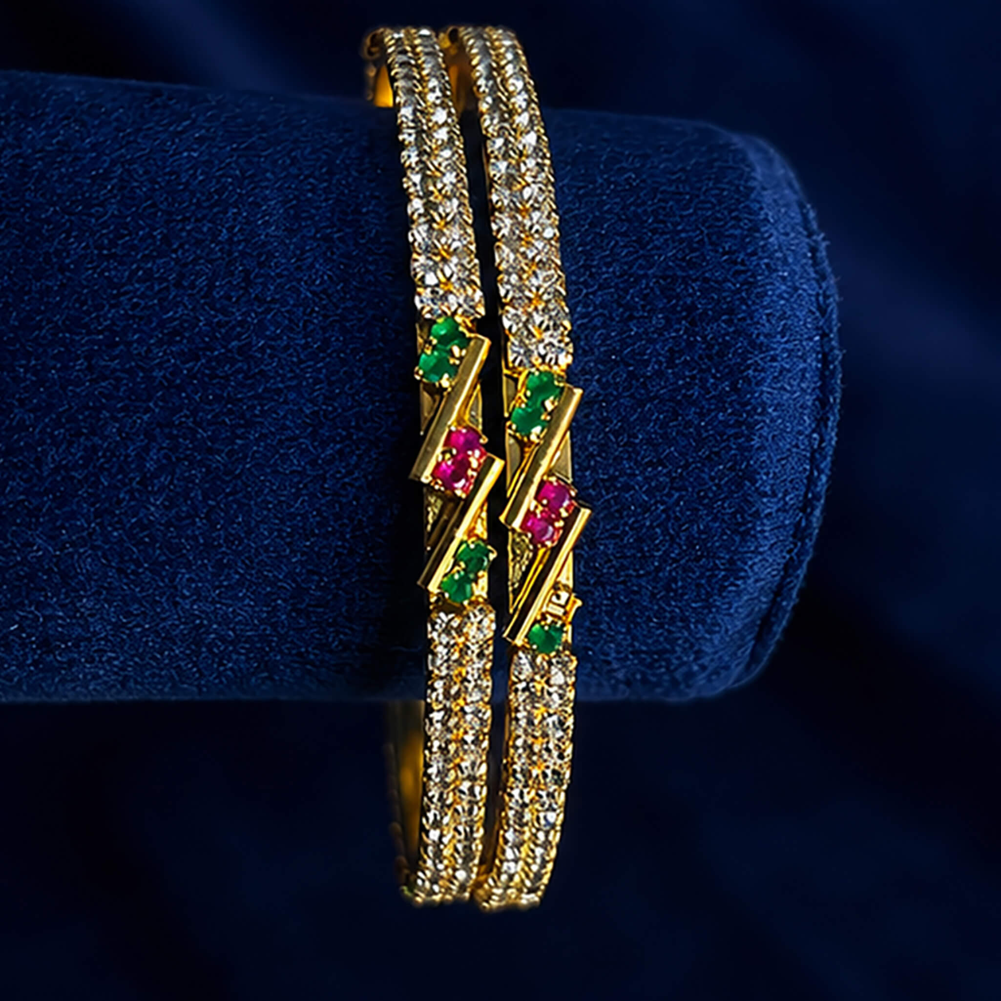 Gold-tone crystal-studded bangles with ruby-pink and emerald-green accents displayed on a blue velvet stand.