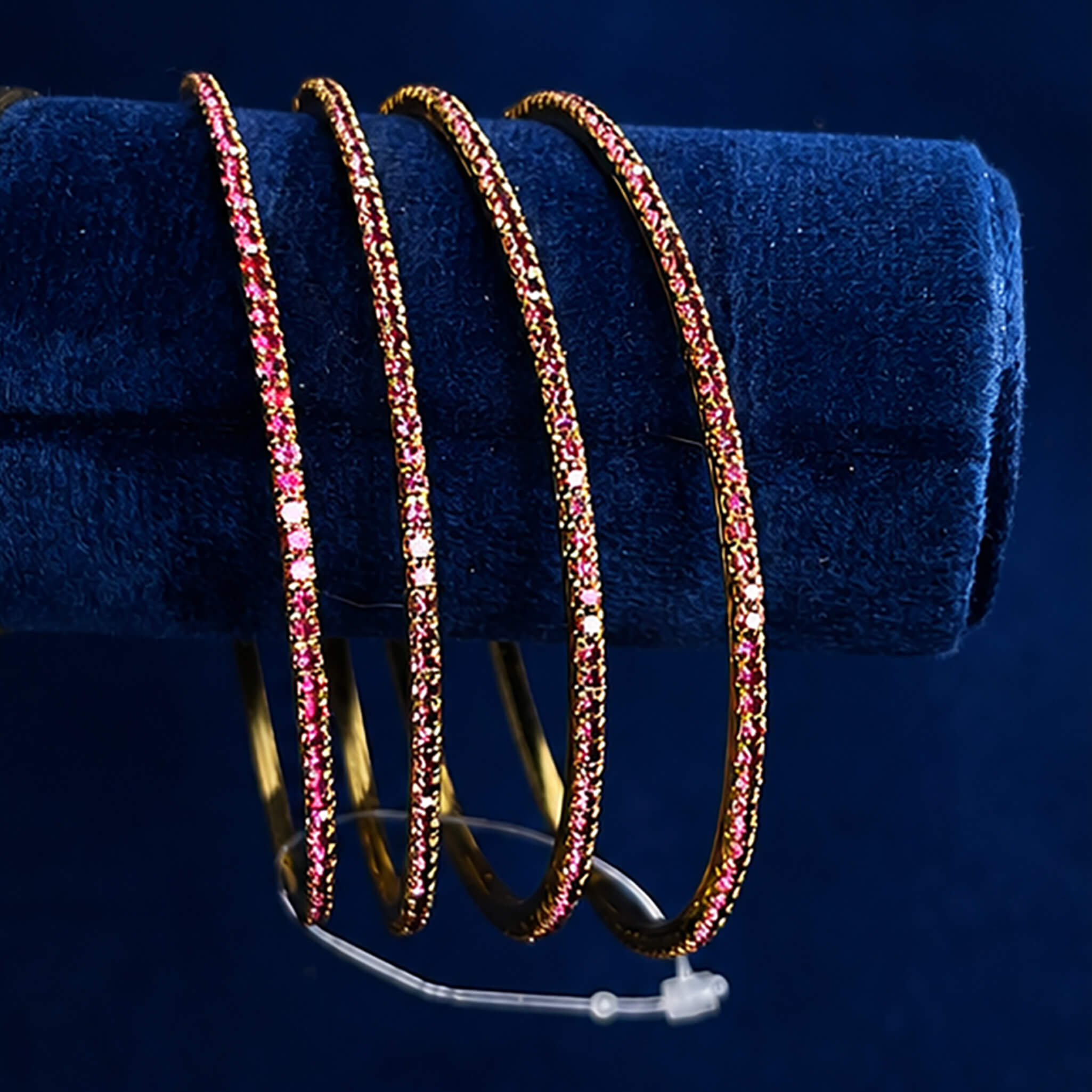 Set of four gold-tone bangles studded with ruby-pink crystals displayed on a blue velvet jewelry stand.