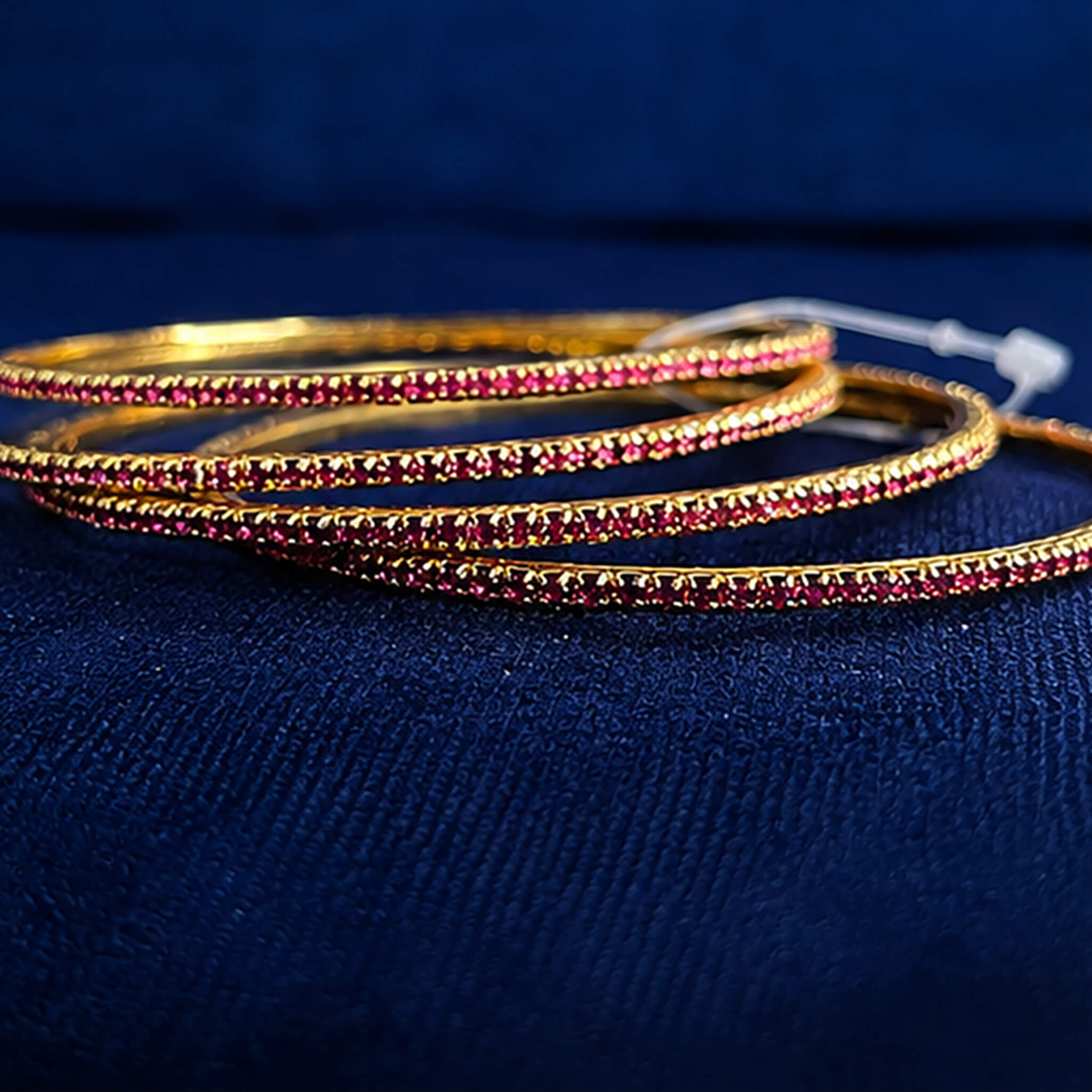 Set of four gold-tone bangles studded with ruby-pink crystals displayed on a blue velvet jewelry stand.