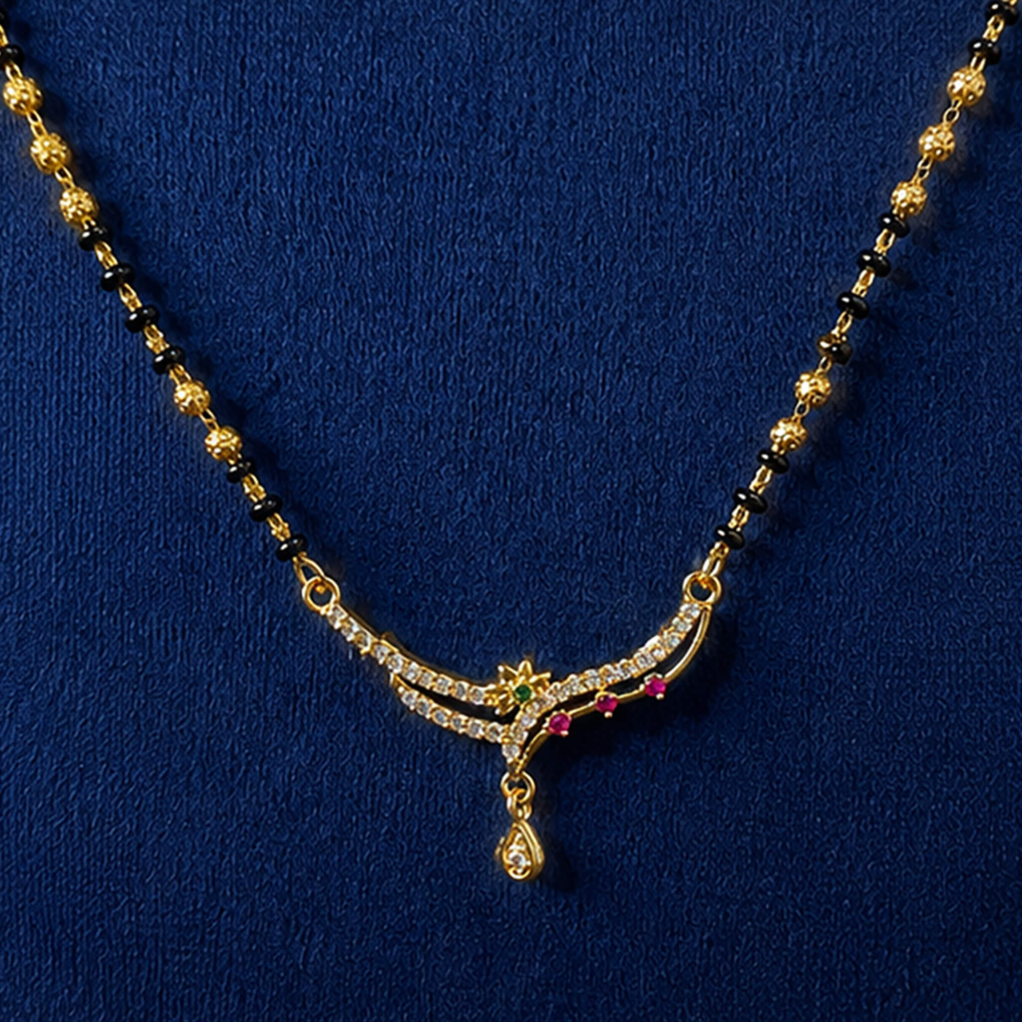 Black-bead mangalsutra necklace with ruby-red stone pendant and dangling charms displayed on a royal blue velvet jewelry bust.