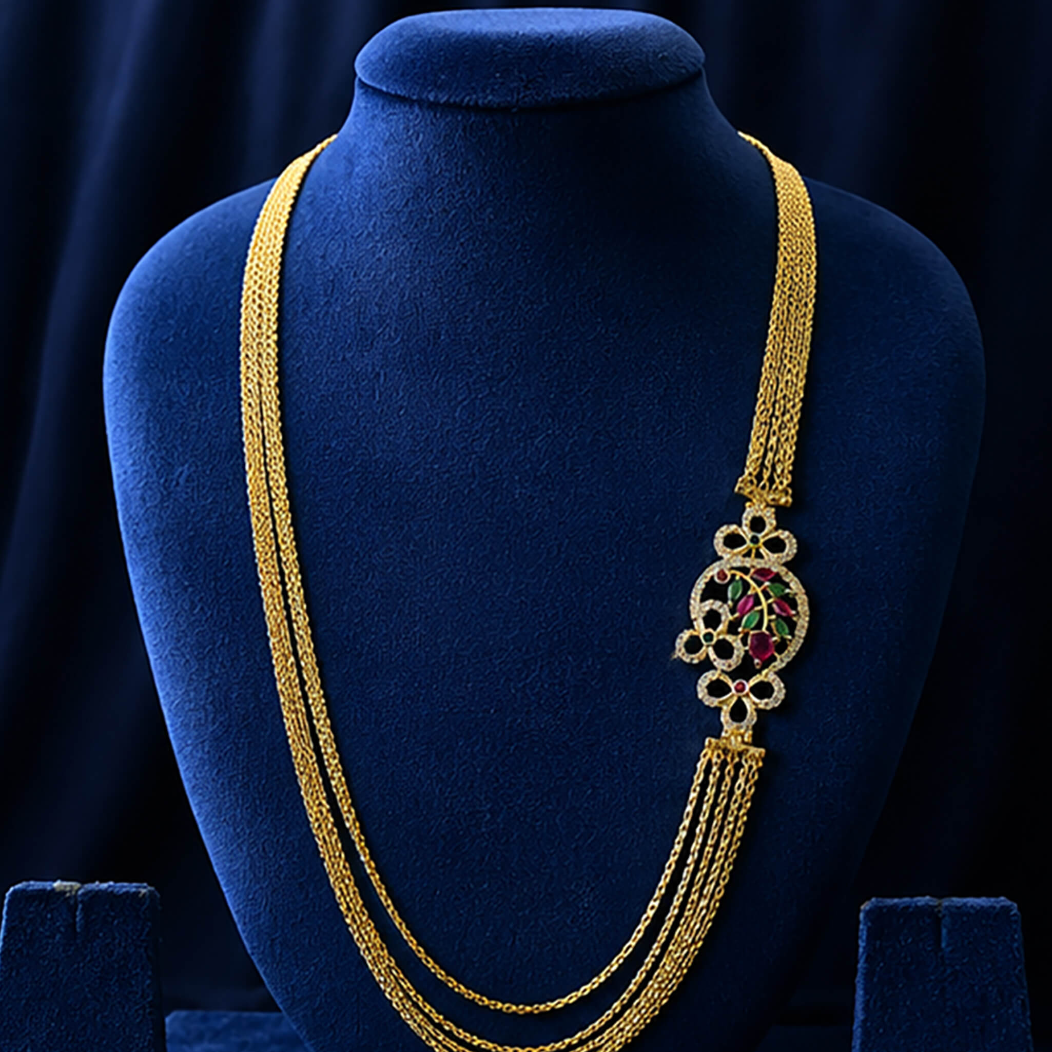 Multi-layered gold-tone Chandraharam with floral vine motif and ruby-green stone accents displayed on a royal blue velvet jewelry bust.