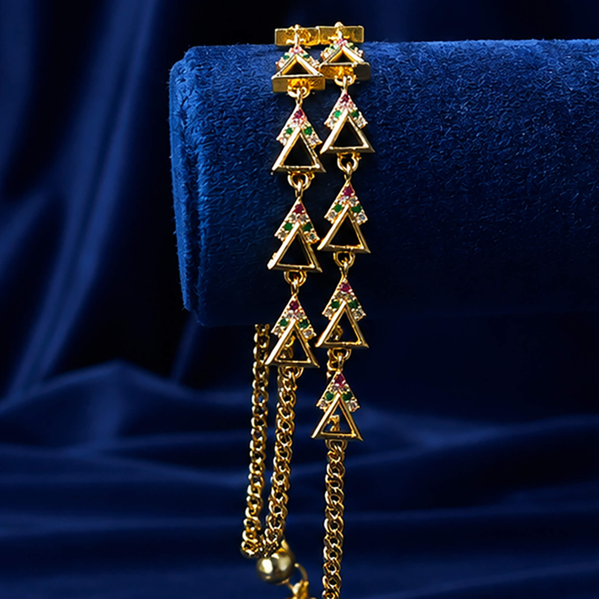 Double-layer gold-tone anklet with geometric triangle charms and colorful crystal accents displayed on a royal blue velvet background.