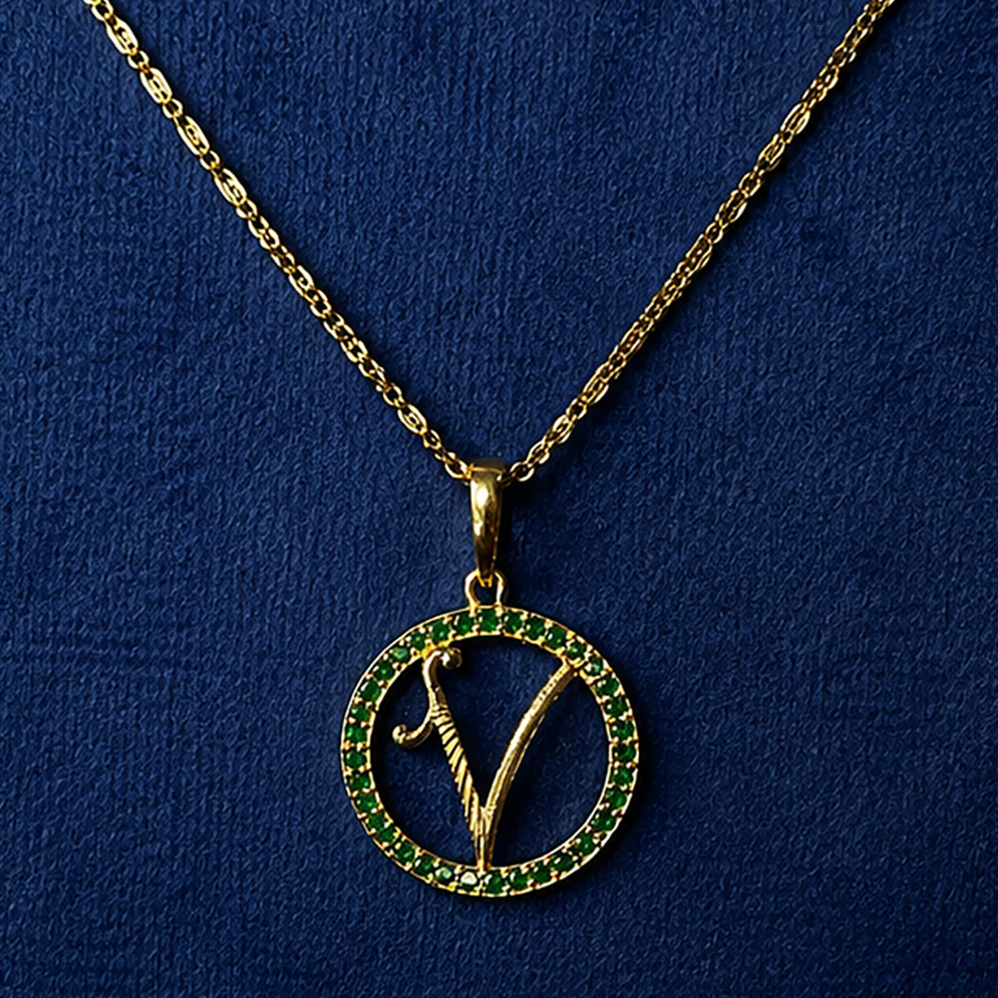 Gold-tone initial V pendant with emerald-green stone accents displayed on a royal blue velvet background.