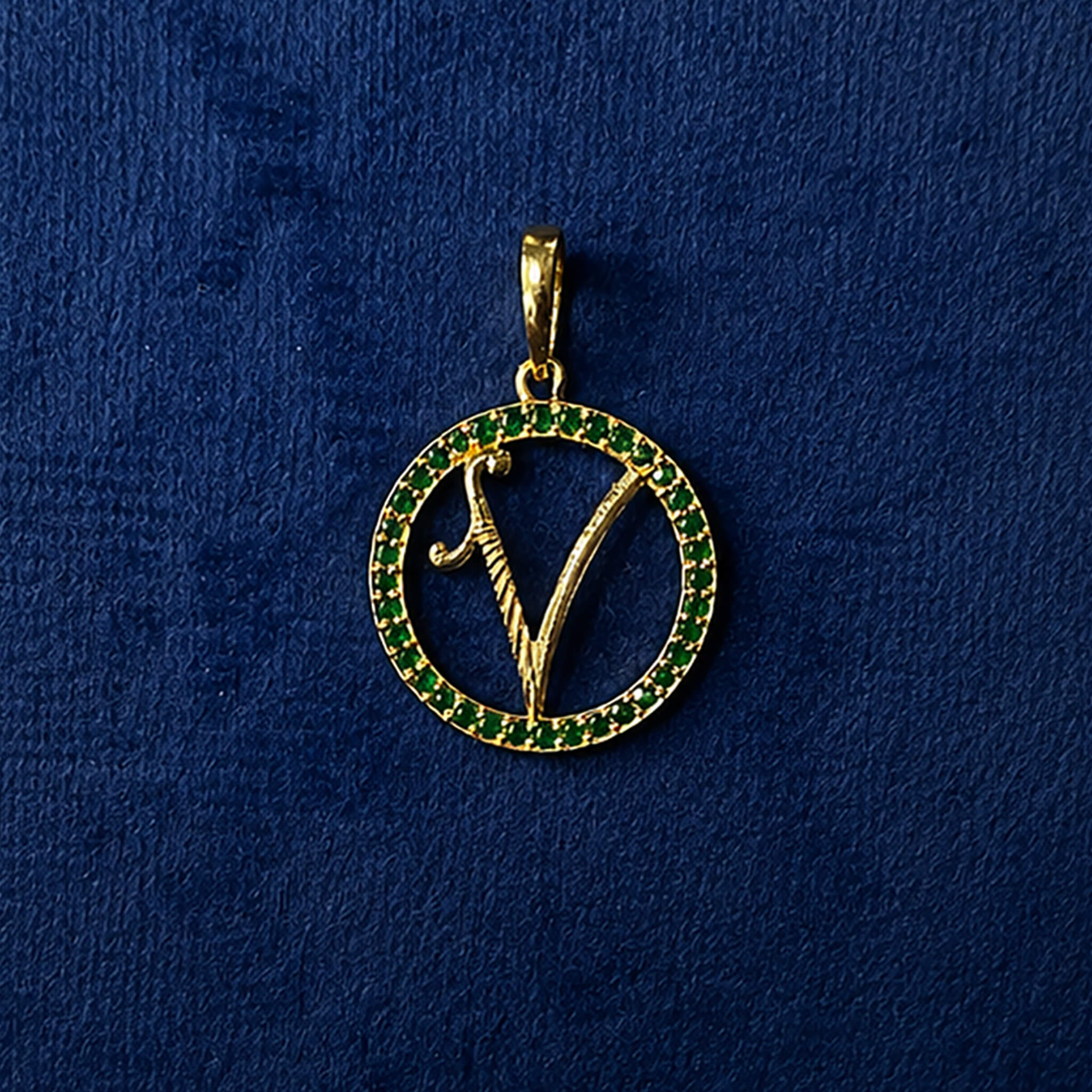 Gold-tone initial V pendant with emerald-green stone accents displayed on a royal blue velvet background.
