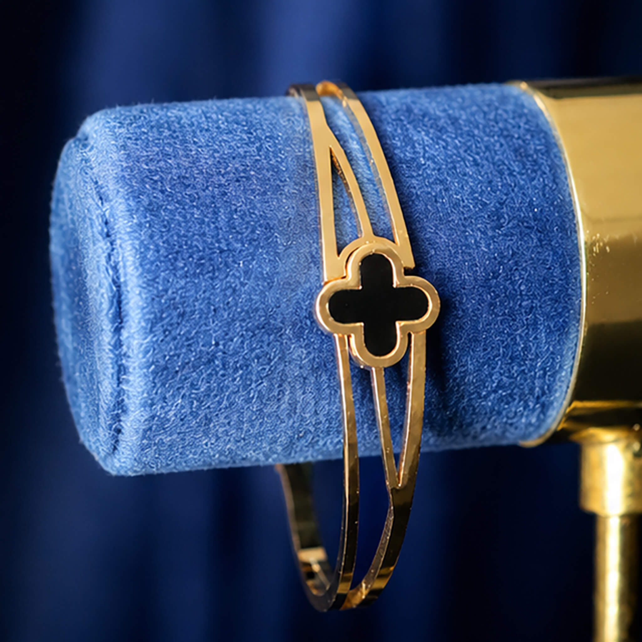 Gold-tone bracelet with a black clover centerpiece displayed on a blue velvet jewelry stand.