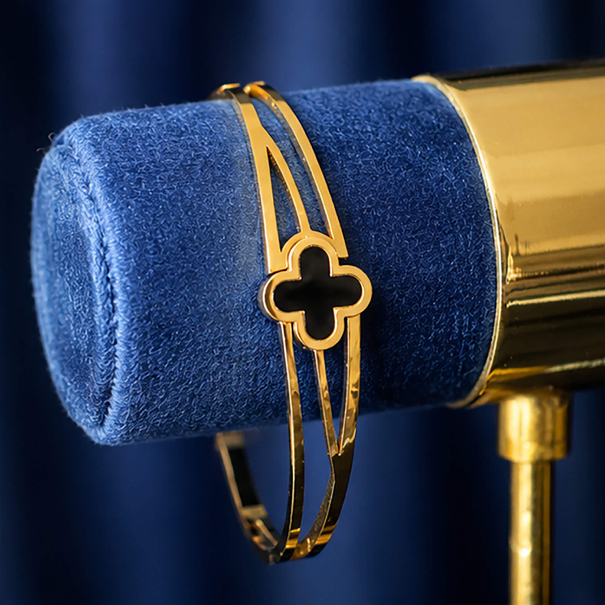 Gold-tone bracelet with a black clover centerpiece displayed on a blue velvet jewelry stand.