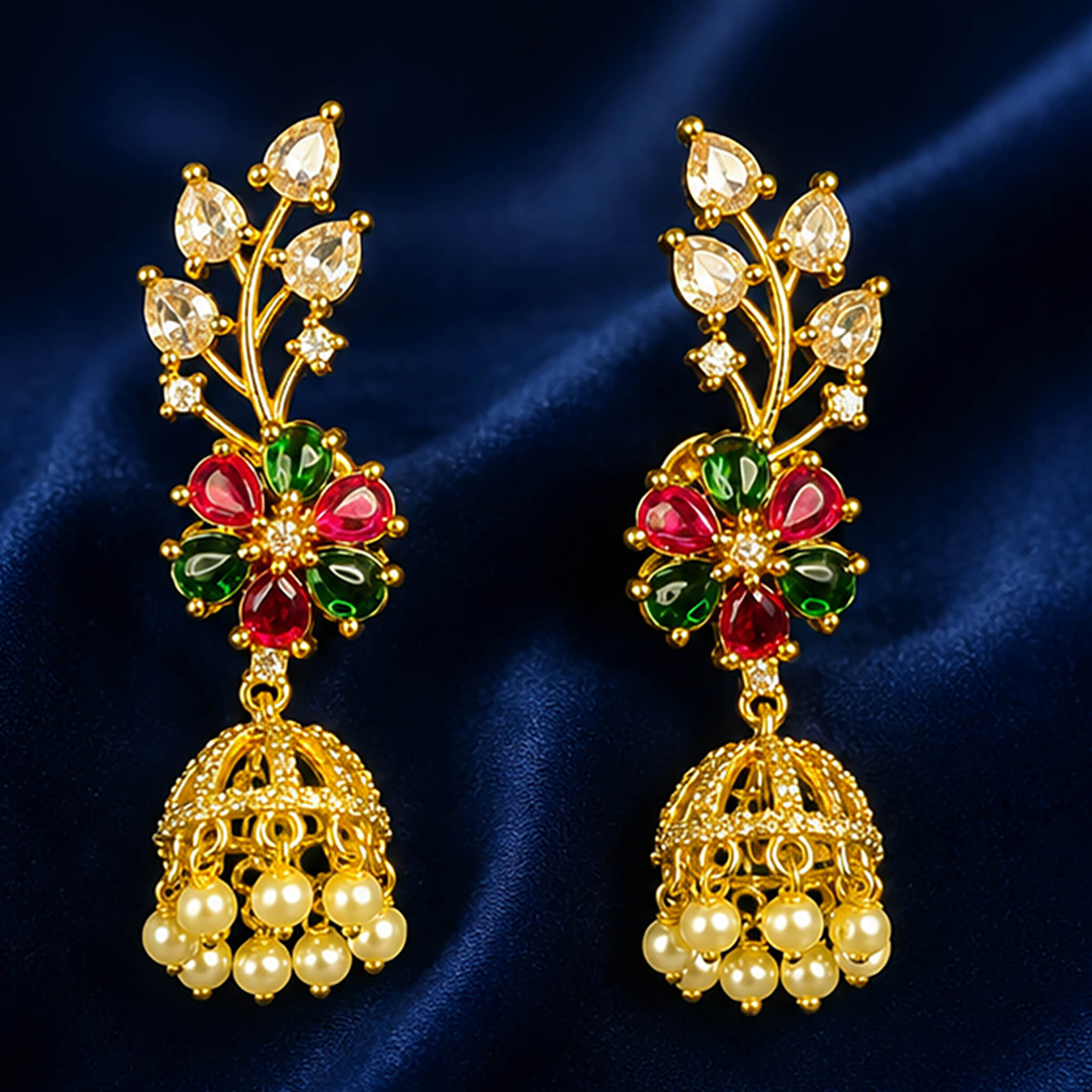 Gold-tone floral jhumka earrings with ruby-red and emerald-green stones and pearl drops displayed on a royal blue velvet stand.