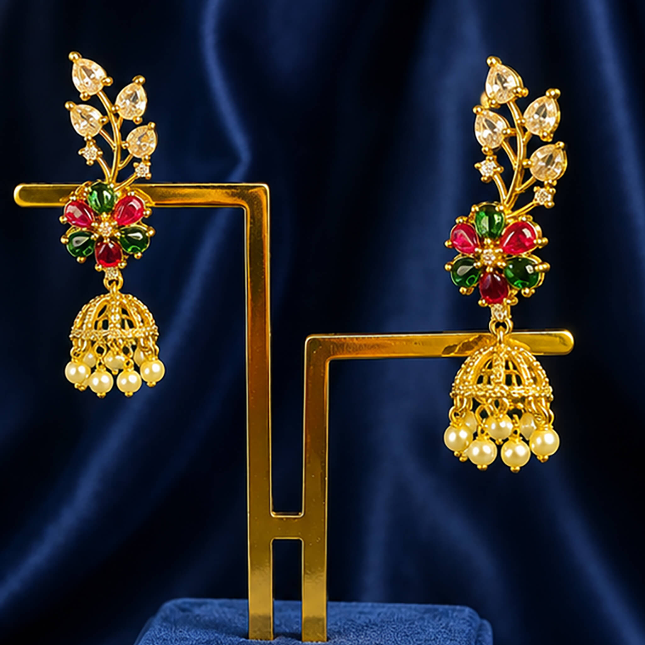 Gold-tone floral jhumka earrings with ruby-red and emerald-green stones and pearl drops displayed on a royal blue velvet stand.