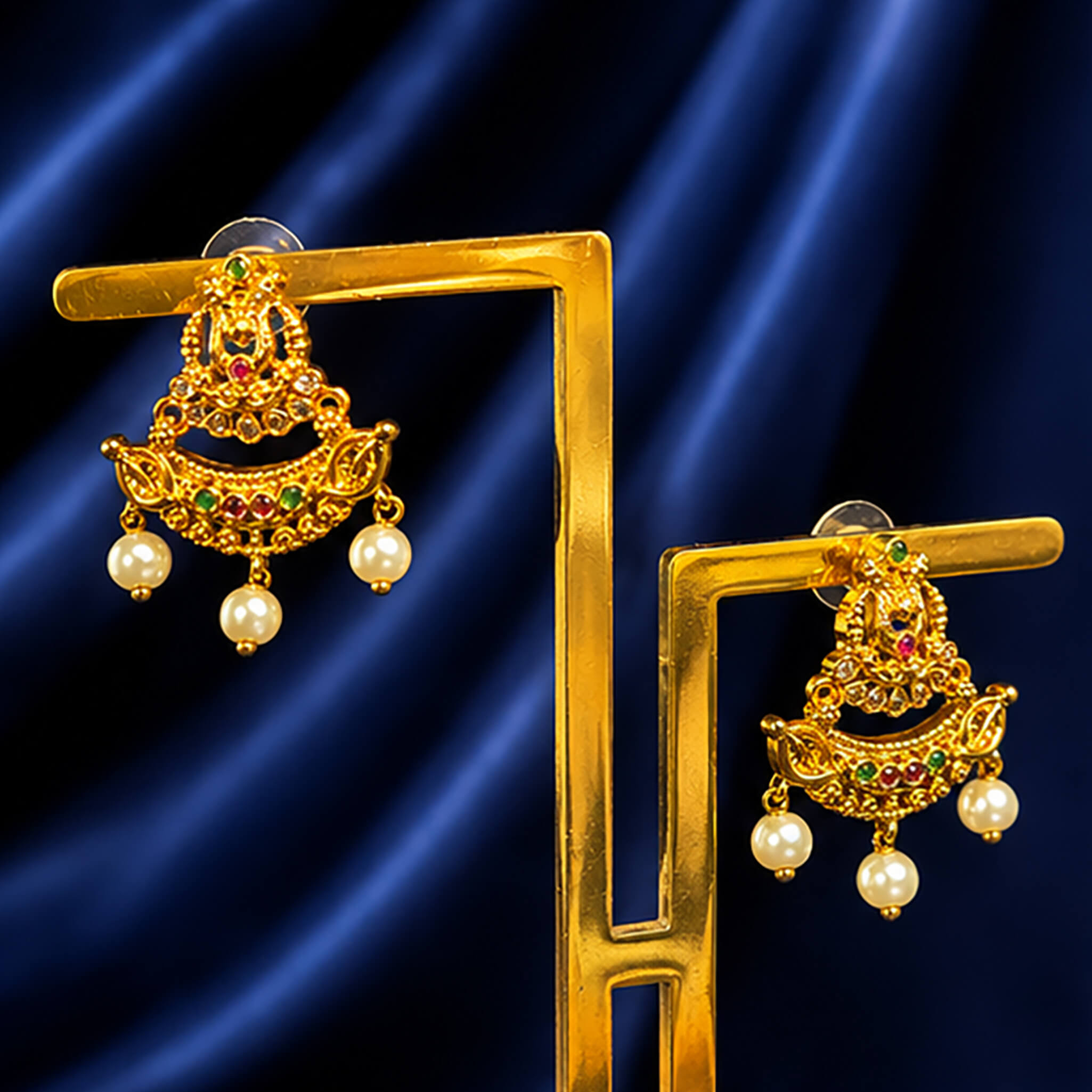 Traditional gold-tone chandbali ear tops with ruby and emerald stones, crystal accents, and pearl drop embellishments on a royal blue background.