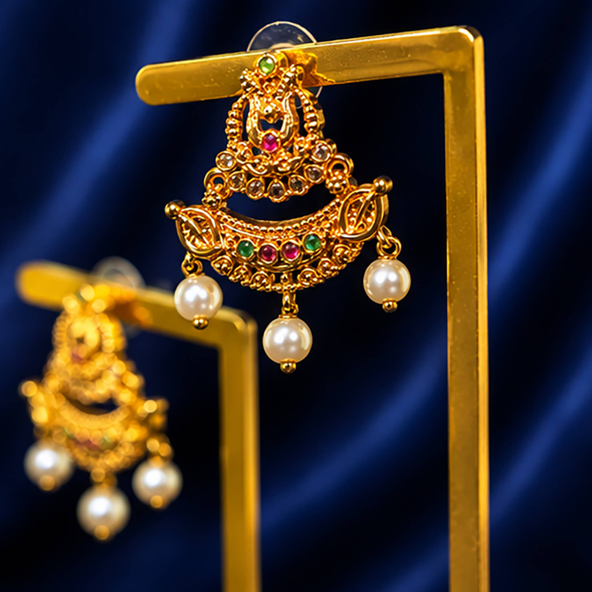 Traditional gold-tone chandbali ear tops with ruby and emerald stones, crystal accents, and pearl drop embellishments on a royal blue background.