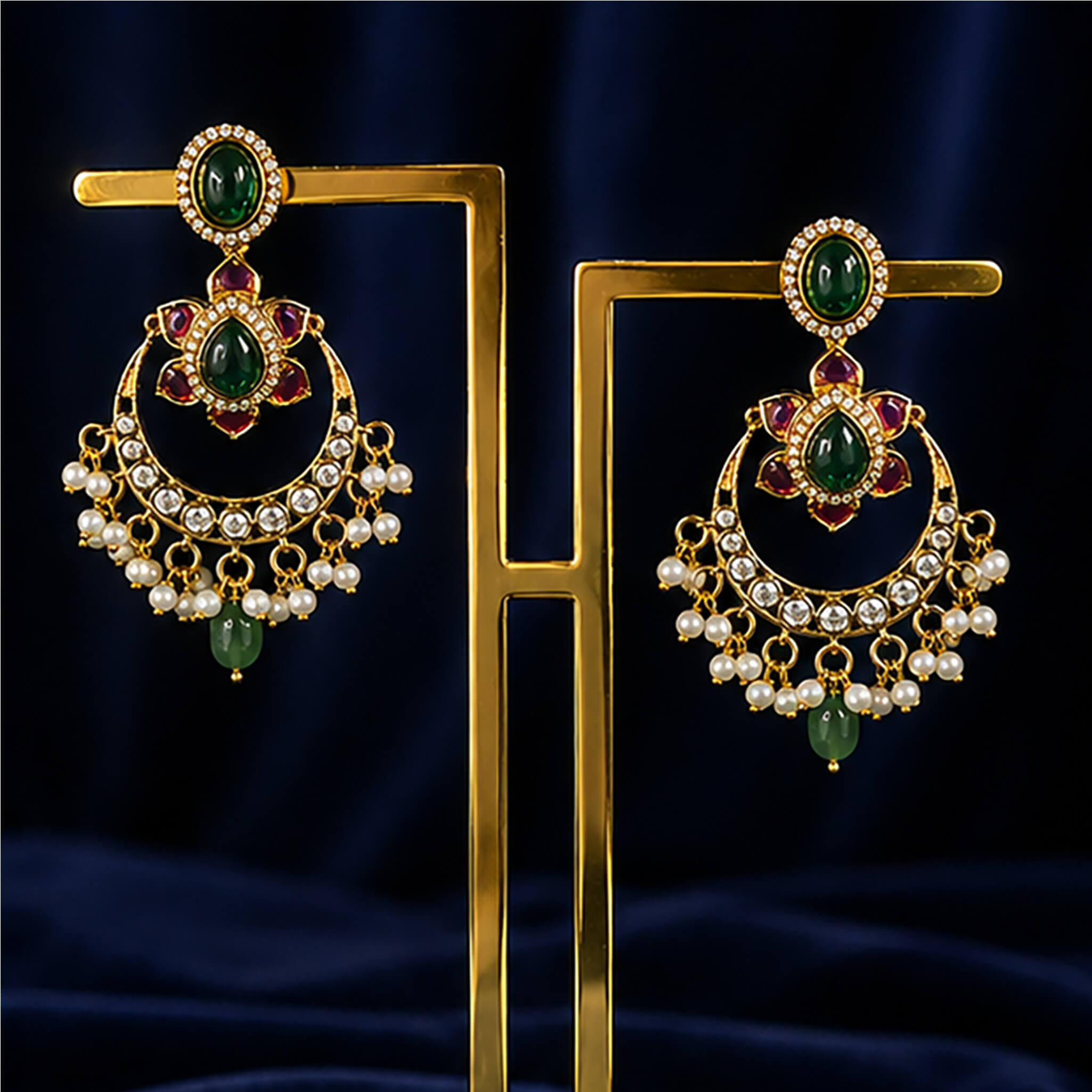 Gold-tone Chandbali earrings with emerald-green stones, ruby accents, crystal detailing, and pearl drops displayed on a luxury jewelry stand.